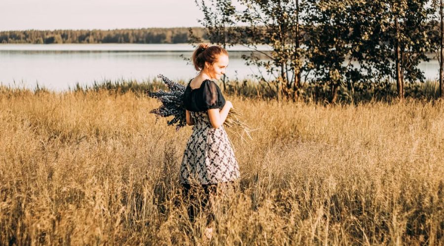 A young woman stands in a sunlit field by a lake, holding lavender flowers and smiling.